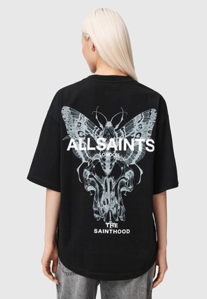 Blond persoon draagt een zwart oversized T-shirt met een grote grijze mot en schedel afbeelding en witte tekst "ALLSAINTS LONDON THE SAINTHOOD" op de achterkant.