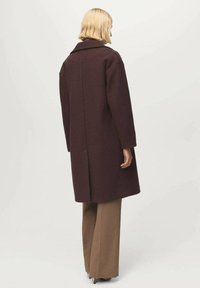 Cappotto di lana borgogna con colletto classico, maniche lunghe, taglio dritto e tasche laterali, abbinato a pantaloni a gamba larga marroni.