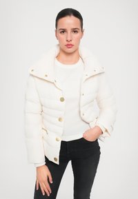 Giacca imbottita bianca con colletto alto, bottoni a pressione dorati e dettagli con zip. Il tessuto ha una texture liscia e un motivo trapuntato.