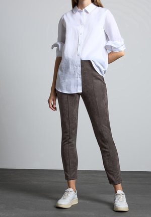 Camicia bianca in lino con maniche arrotolate, abbinata a pantaloni aderenti in suede marrone con cuciture a vista e sneakers bianche.