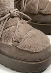 Brown footwear featuring a soft, fuzzy upper with textured fabric and a sturdy, ridged sole. Laces are integrated into the design.