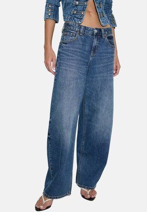 Person iført viddeblå denimjeans, kort denimjakke og hvide tåsandaler, stående mod en ensfarvet baggrund.
