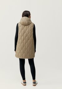 Beige vadderad ärmlös väst med huva, med en slät textur och rundade mönster, kombinerad med svarta leggings och lätta skor.