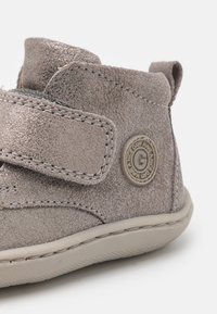 Bota infantil de ante gris con una correa de velcro, puntera redondeada y suela de goma texturizada gris. Presenta un logo redondo en el lateral.