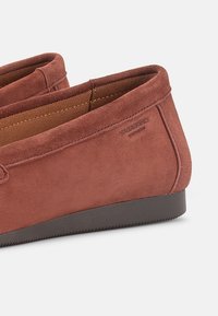 Κοντινό πλάνο καφέ σουέντ παπουτσιών χωρίς κορδόνια με μαύρη σόλα και λεπτή ραφή, που δείχνει την εσωτερική επένδυση και την ανάγλυφη σήμανση της μάρκας.