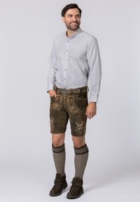 Graues Langarmhemd mit Knöpfen, braune Leder-Shorts mit Stickerei, graue kniehohe Socken und dunkle Stiefel, vor einem hellen Hintergrund stehend.