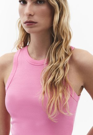 Young woman with long wavy blonde hair wearing a sleeveless pink ribbed top, standing against a plain white background.