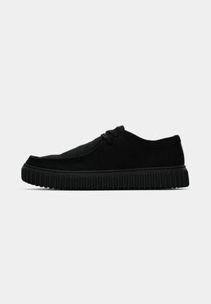 Black low-top casual shoe with rounded toe, lace-up front, suede-like texture, and thick ridged sole, shown in side profile on white background.