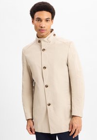 Beige trenchcoat met een hoge kraag, grote knopen, zijzakken en een gladde textuur, op maat gemaakt voor een getailleerde uitstraling.
