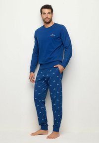 Blauer Baumwollpullover mit rundem Halsausschnitt. Passende blaue Hose mit kleinem Auto-Muster; elastische Bündchen und Bund. Model steht barfuß.