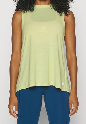 Vrouw draagt mouwloos lichtgroen sporttopje en donkerblauwe legging, staand met armen ontspannen langs haar lichaam.