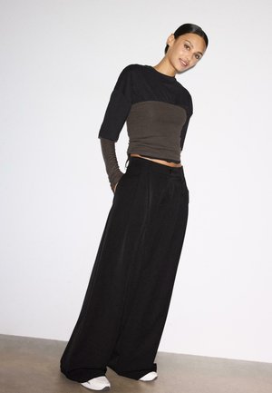 Žena s tmavými vlasmi v nízkom drdole, oblečená v vrstvených čiernych a hnedých topoch, širokých čiernych nohaviciach a bielych teniskách stojaca pri bielej stene.