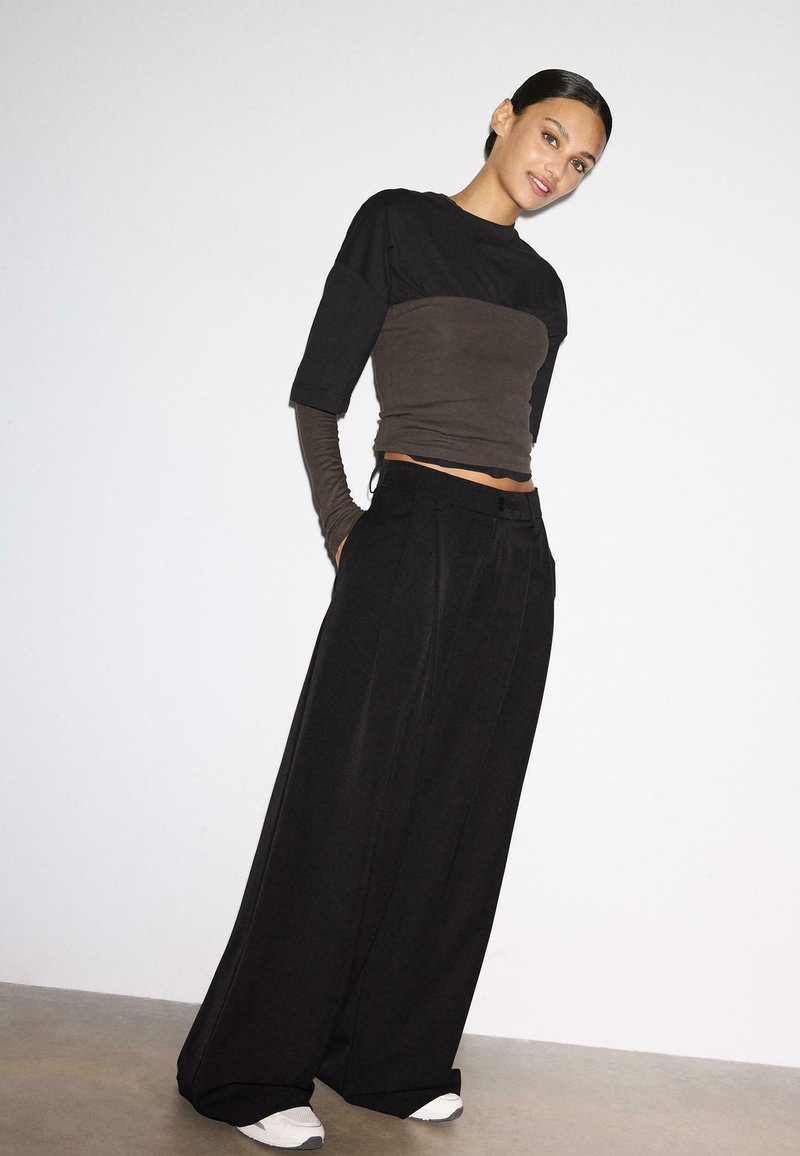 Femme aux cheveux foncés coiffés en chignon bas, portant des hauts superposés noirs et marron, un pantalon large noir, et des baskets blanches, se tenant contre un mur blanc.