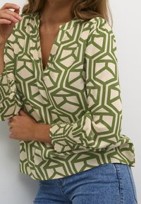 Blouse à motif géométrique vert et crème avec un col en V, des poignets smockés et une coupe décontractée, associée à un jean en denim.