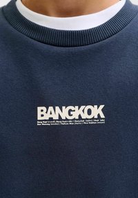 Primo piano di una felpa girocollo blu navy con scritta bianca "BANGKOK" e nomi dei distretti più piccoli stampati sotto sul petto.