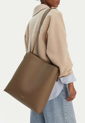 Kobieta w beżowym płaszczu i jeansowej spódnicy, niosąca dużą brązową skórzaną torbę na ramię z złotym suwakiem i logo.