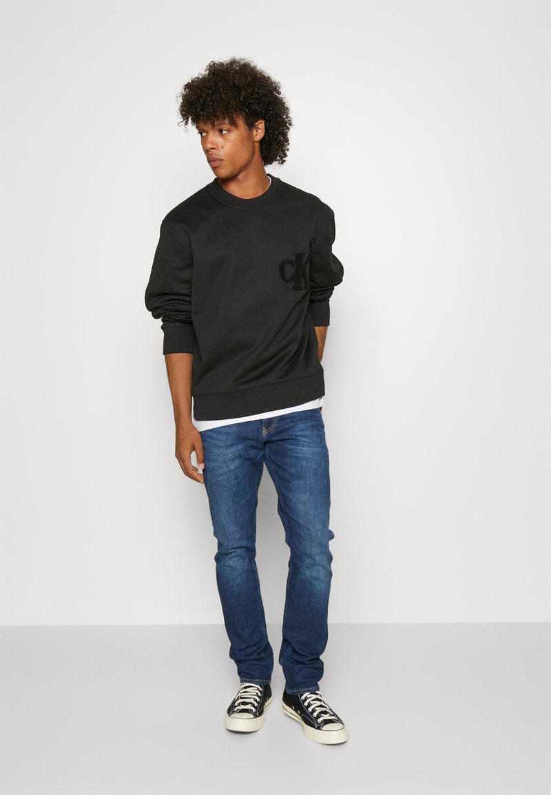 Black sweatshirt with a front embossed logo, round neck, and ribbed cuffs paired with blue jeans and black sneakers with white soles.