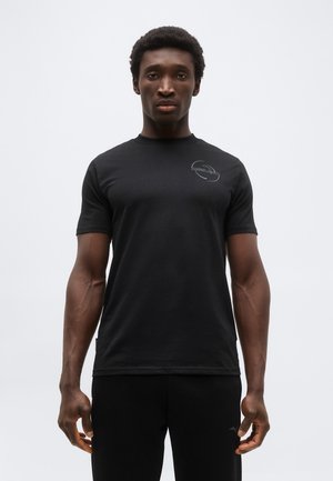 Młody mężczyzna stojący, ubrany w dopasowaną czarną koszulkę i czarne spodnie, na jednolitym jasnoszarym tle.