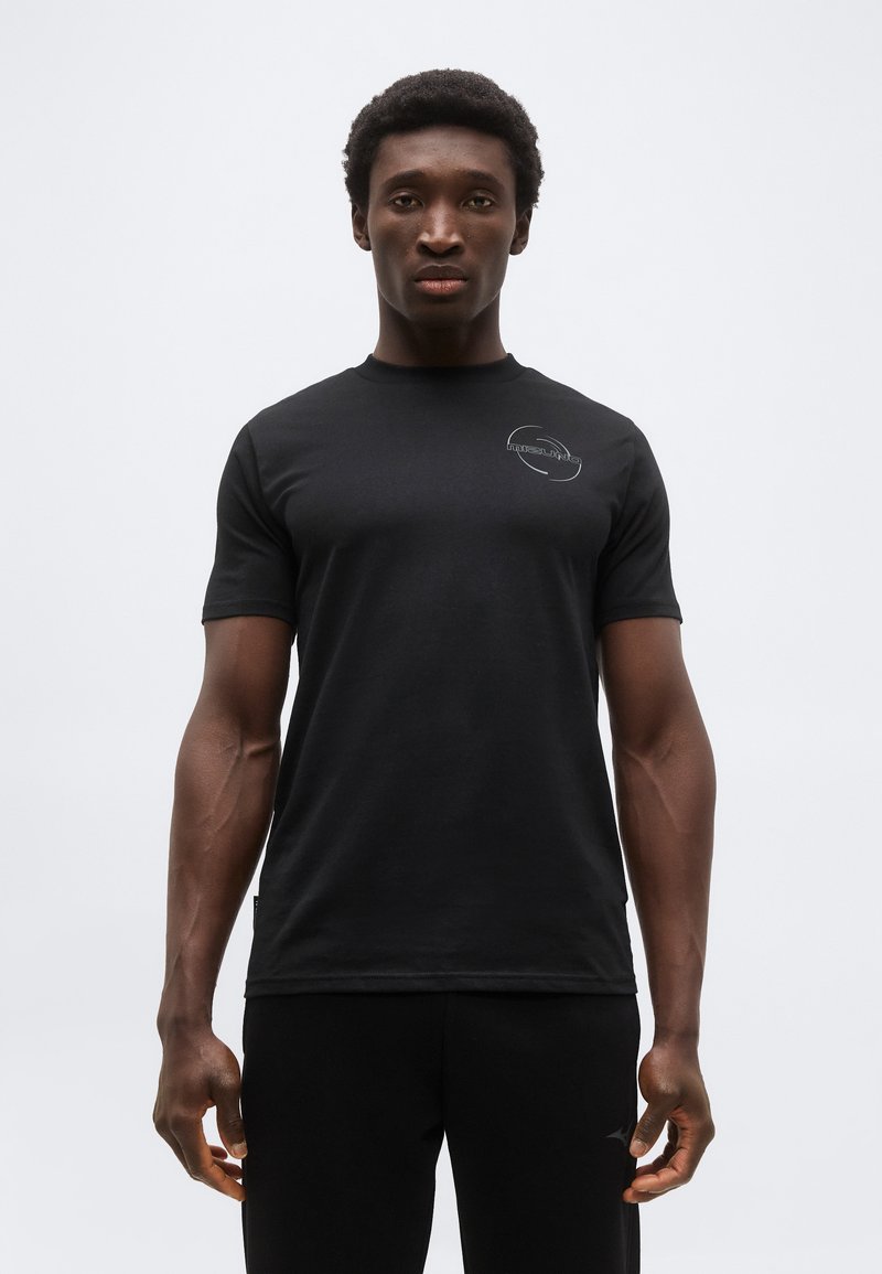 Νεαρός άνδρας όρθιος, φοράει εφαρμοστή μαύρη μπλούζα και μαύρο παντελόνι, μπροστά σε απλό ανοιχτό γκρι φόντο.