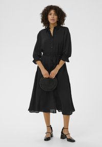 Czarna, długa sukienka z długim rękawem, z przodu zapinana na guziki, z paskiem w talii i falbaniastymi mankietami. Połączona z plecioną czarną okrągłą torebką i butami na obcasie.