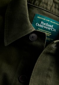 Tumši zaļš auduma krekla apkakle ar pogu un birku no Portland Outerwear Co., kurā redzami zīmola dati un rūpes par apģērbu.