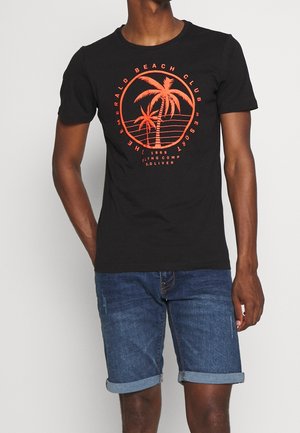 Homme portant un t-shirt noir avec un dessin de palmier orange et du texte, associé à un short en denim bleu retroussé, debout devant un fond blanc.