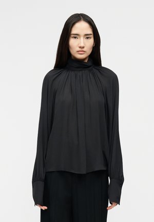 Γυναίκα με μακριά ίσια μαλλιά, που φοράει μια φαρδιά μαύρη μπλούζα με ψηλό λαιμό, με σούρες στο ύφασμα και φαρδιές μανσέτες, στέκεται μπροστά σε ένα απλό φόντο.