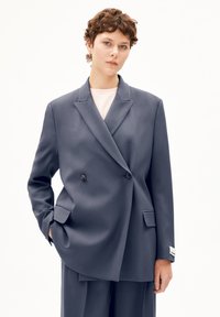 Blazer doppiopetto in blu scuro, realizzato in tessuto liscio con colletto a patta, tasche laterali e un'etichetta bianca sul polsino.
