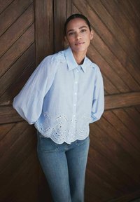 Kvinde med mørkt hår iført en blå stribet bluse med hulmønsterkant og jeans står foran en væg beklædt med træpaneler og ser ind i kameraet.