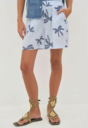 Žena u plavim i bijelim prugastim kratkim hlačama s printom palmi i zlatnim sandalama gladiatorskog stila s nitnama, stoji s jednom rukom u džepu.