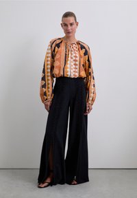 Multikleurige blouse in oranje, zwart en wit met een striknek, gecombineerd met zwarte wijde broek met zijslits en textuurdetails.