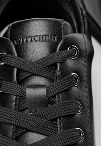 Sneaker in pelle nera in primo piano con superficie textured, lacci neri rotondi, occhielli metallici e branding "WITCHE" impresso sulla linguetta.