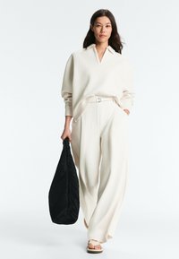 Femme portant une chemise blanche ample et un pantalon large, tenant un grand sac noir texturé, marchant vers l'avant sur fond blanc.
