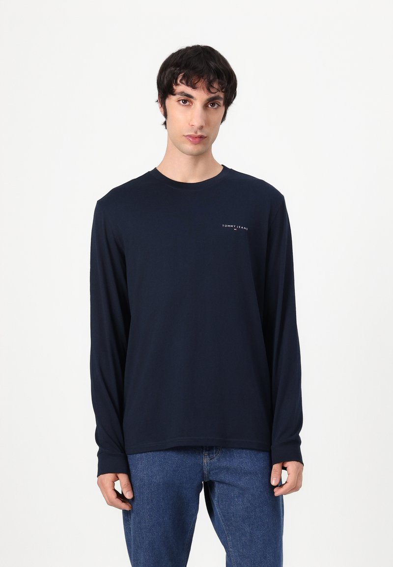 Marineblå langermet T-skjorte laget av myk bomull, med rund hals og subtil logo på brystet; kombinert med blå denim jeans.