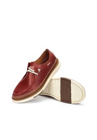 Des chaussures slip-on en cuir rouge avec une semelle crème mouchetée, présentant un design cousu et un accent de lacet décoratif sur le côté.