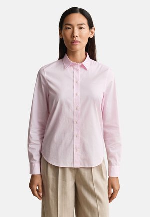 Žena v svetloružovej košeli na gombíky a béžových plisovaných nohaviciach, pozerajúca sa vpred s neutrálnym výrazom na jednoduchom pozadí.