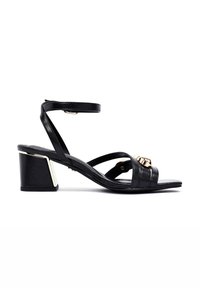 Sandalias de tacón en bloque de cuero negro con tiras entrelazadas, correa de tobillo ajustable y detalles en tono dorado en la parte delantera y en los acentos del tacón.