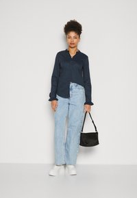 Námořnická košile s knoflíky a volánkovými manžetami, světle modré džíny rovného střihu, bílé platformové tenisky a malá černá crossbody kabelka.