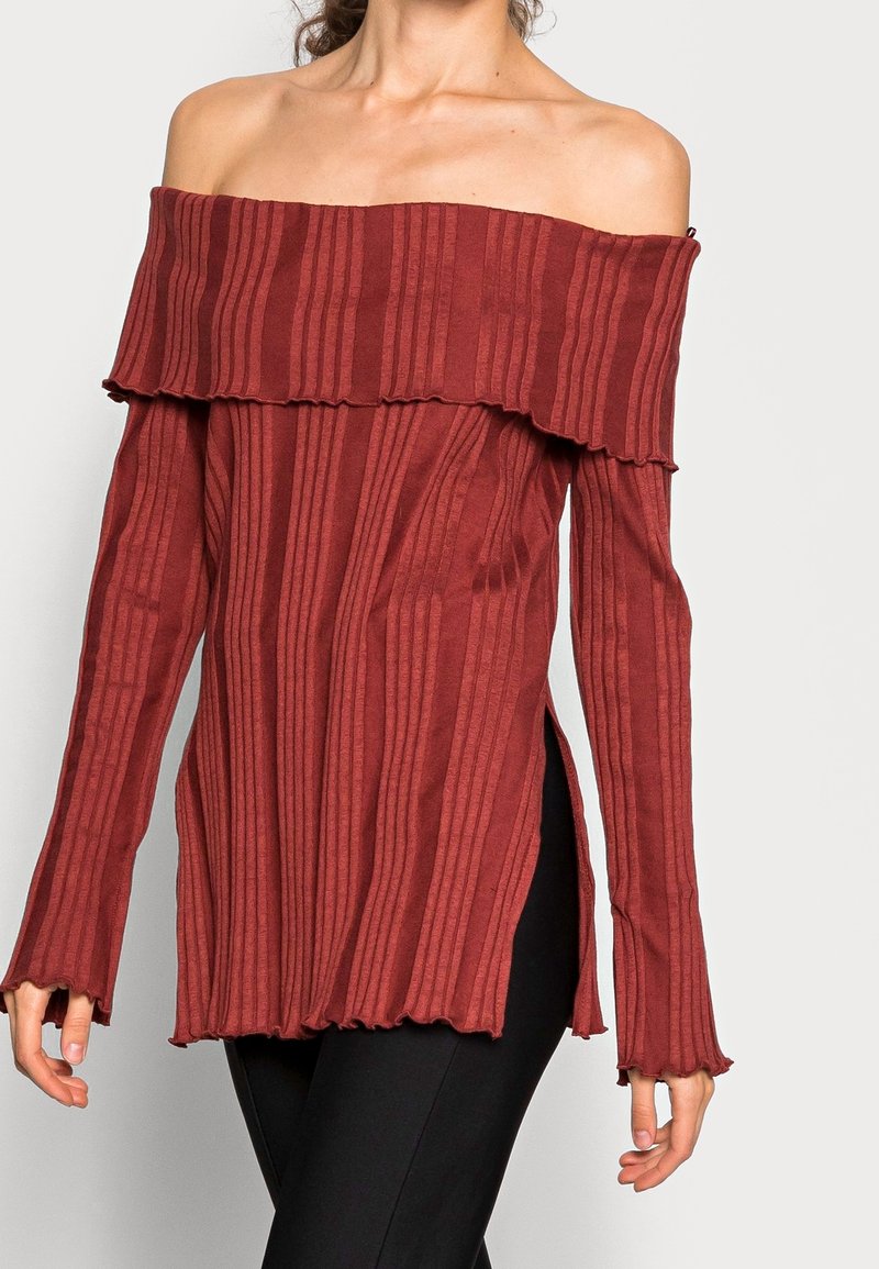 Off-the-shoulder, ribbed top in rust red with flared sleeves, side slits, and wavy hem details, crafted from soft, stretchy material.