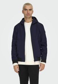 Giacca zip navy con cappuccio, tasche frontali e polsini neri. Indossata sopra una felpa bianca e pantaloni neri. Tessuto a superficie liscia.