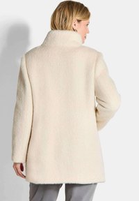 Crème-kleurige jas met een gestructureerde afwerking, voorzien van een hoge kraag en lange mouwen, met een zoom die onder de heupen valt, eenvoudig ontwerp zonder zichtbare sluitingen.