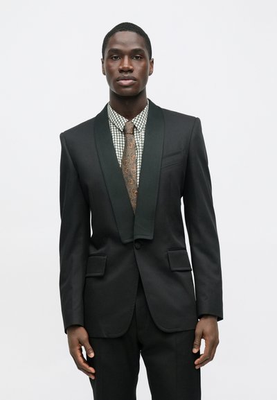 Čierny smokingový kabát s zeleným saténovým šálovým golierom, doplnený o károvanú košeľu a hnedú kravatu s paisley vzorom. Viditeľné sú dve predné vrecká.