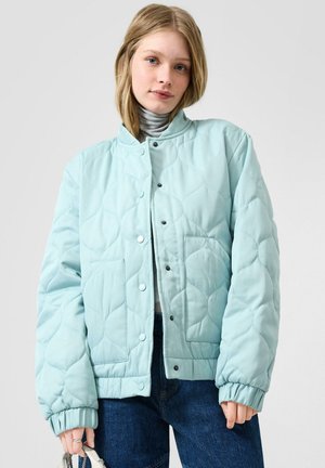 Jonge vrouw met een lichtblauwe gewatteerde jas over een grijze coltrui, gecombineerd met een blauwe spijkerbroek, en een kleine witte tas in haar hand.