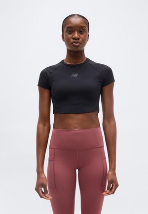 Ung kvinde stående iført en sort cropped atletisk top og højtaljede mauve leggings, vendt fremad mod en ensfarvet baggrund.