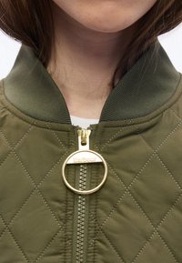 Κοντινό πλάνο του λαιμού και του πηγουνιού ενός ατόμου που φοράει ένα πράσινο ραμμένο μπουφάν με μεγάλο κυκλικό χρυσό φερμουάρ που γράφει "Barbour".