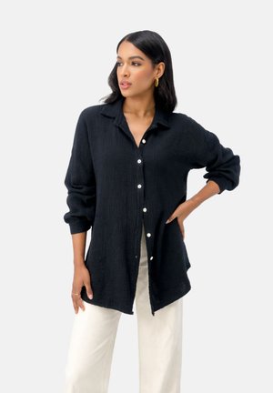 Frau mit langen dunklen Haaren, die ein lockeres schwarzes Hemd mit Knöpfen und weiße Hose trägt, steht mit einer Hand in der Hüfte vor einem schlichten Hintergrund.