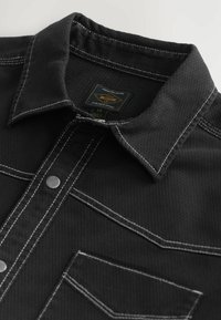 Black button-up shirt made of textured fabric. Features white stitching, a pointed collar, and two front pockets with a snap closure.