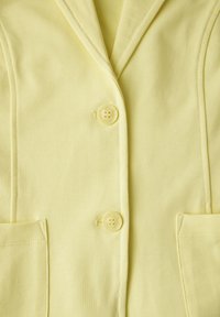 Geel jasje met een reverskraag met inkeping, twee knopen en zijzakken. Gemaakt van zachte stof met een gladde, gelijkmatige textuur.