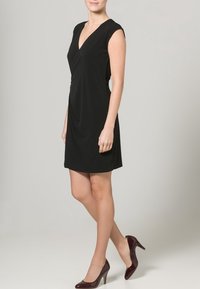 Černé šaty typu shift s krátkými rukávy a výstřihem do V, s volně drapovanou tkaninou a přiléhavým střihem, doplněné burgundskými jehlovými podpatky.