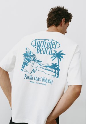 Homme portant un t-shirt blanc avec un motif bleu Surfrider Beach comprenant des palmiers, l'océan, un surfeur et le texte Pacific Coast Highway au dos.